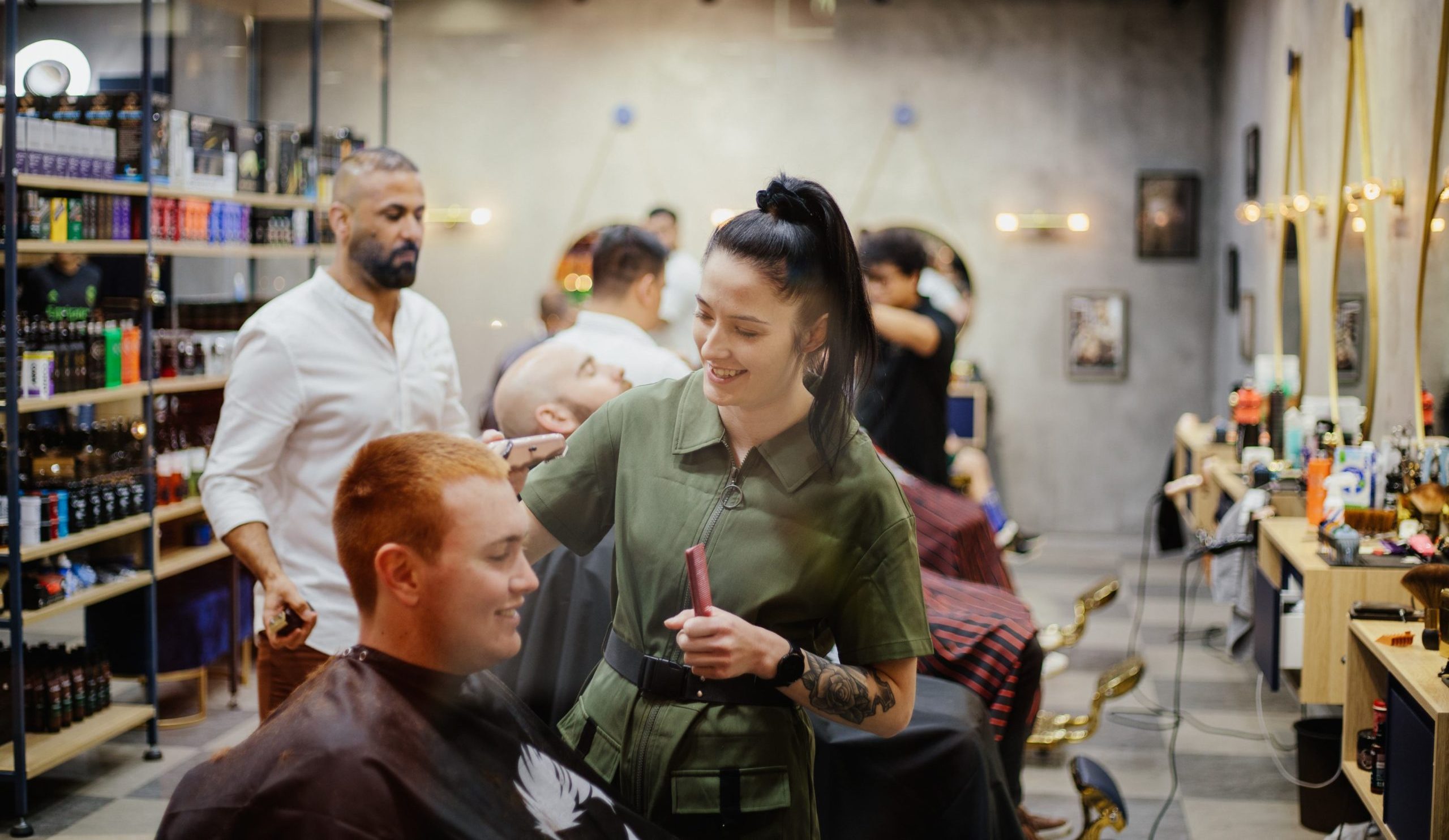 A hairdresser in a green shirt smiles as she cuts the hair of a seated man with reddish hair in a busy barbershop.